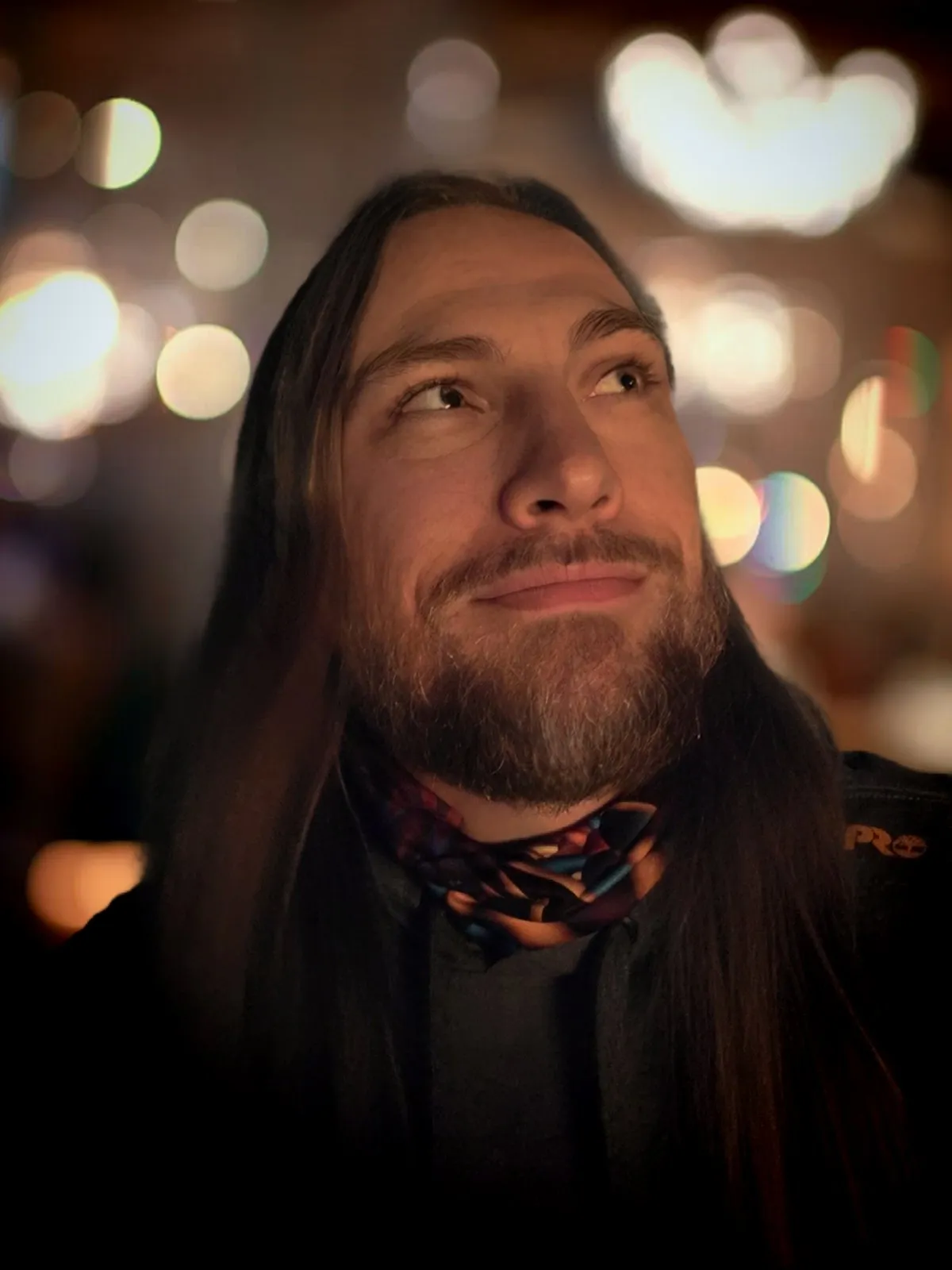 Matt Cheramie, bearded and long-haired, smiling at something off camera in a bar at night, the lights behind him out of focus.
